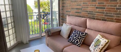 A modern living room with a brick wall, a sofa with patterned cushions, a white table, and a view through a glass door to an outdoor balcony.