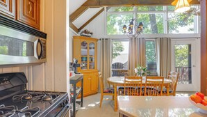 Una cocina con estufa, microondas, armarios de madera, una mesa de comedor con sillas y un ventanal con cortinas.