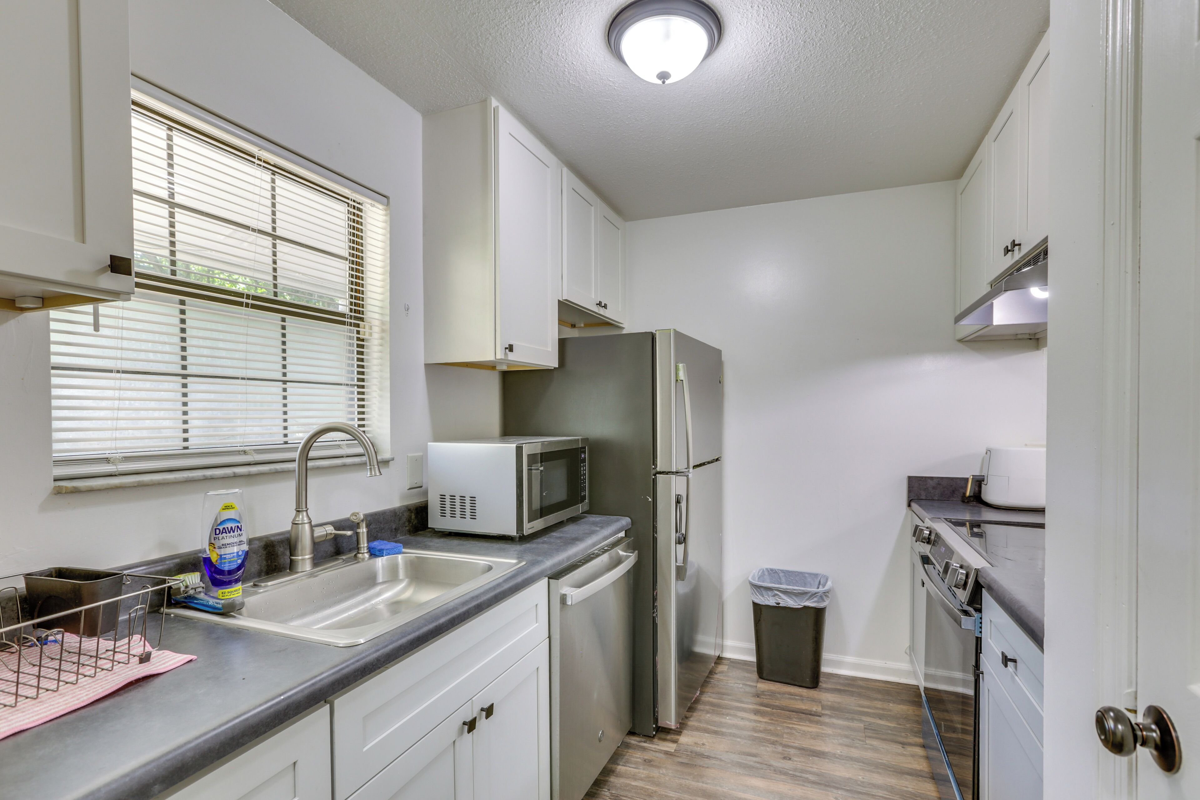 A compact kitchen with white cabinets, a stainless steel refrigerator, a microwave, a sink, and a trash bin.