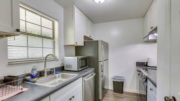 A compact kitchen with white cabinets, a stainless steel refrigerator, a microwave, a sink, and a trash bin.