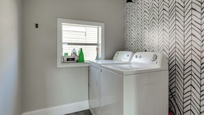 Un cuarto de lavandería con lavadora y secadora, una ventana con útiles de limpieza, y un revestimiento de pared estampado.