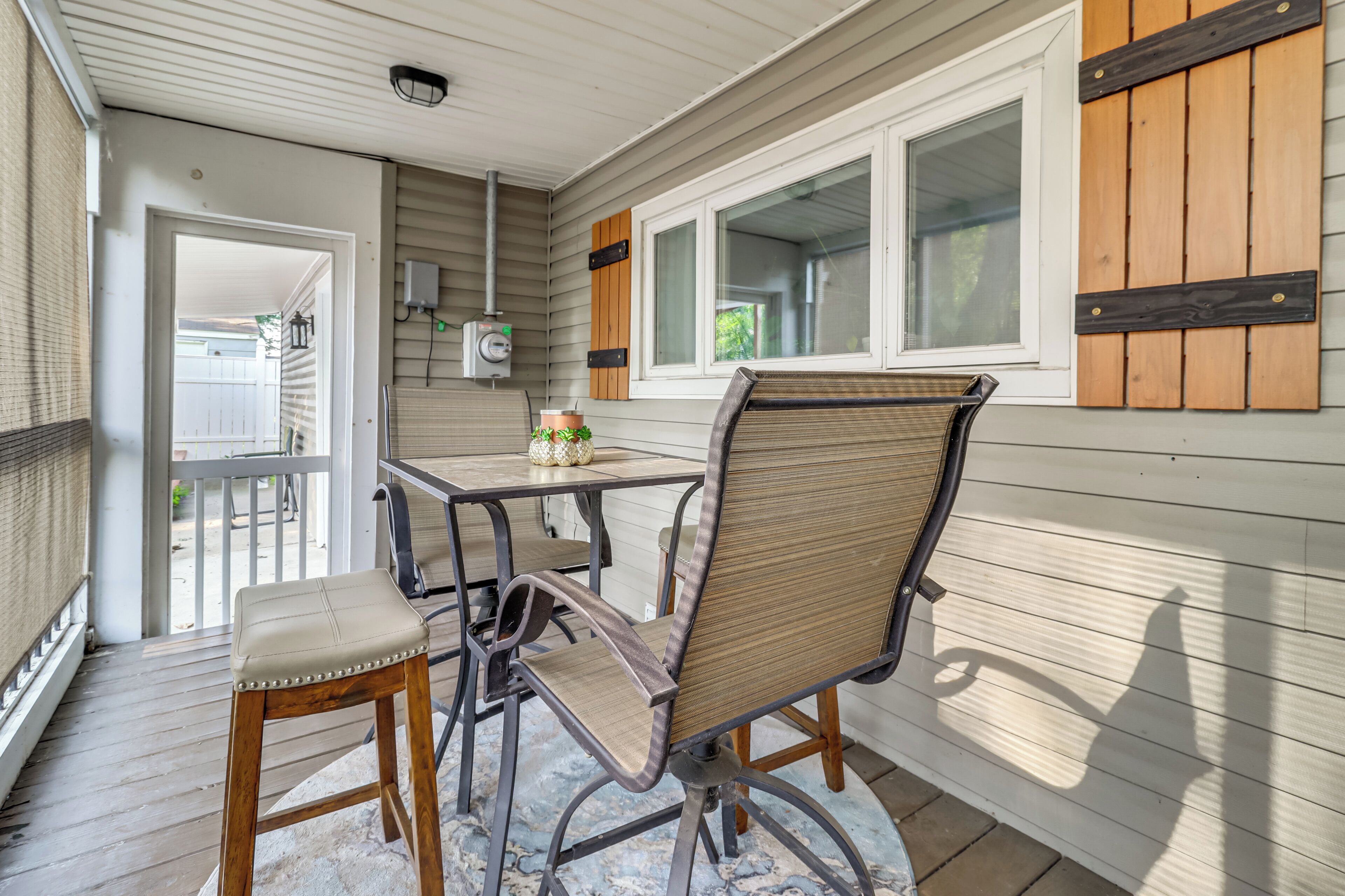 A covered patio area with a table and two chairs, a potted plant, and a window with shutters.