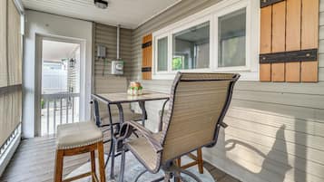 A covered patio area with a table and two chairs, a potted plant, and a window with shutters.