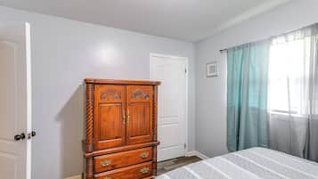 A bedroom with a wooden dresser, a bed with a patterned bedspread, a white door, and a window with curtains.