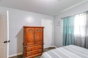 A bedroom with a wooden dresser, a bed with a patterned bedspread, a white door, and a window with curtains.