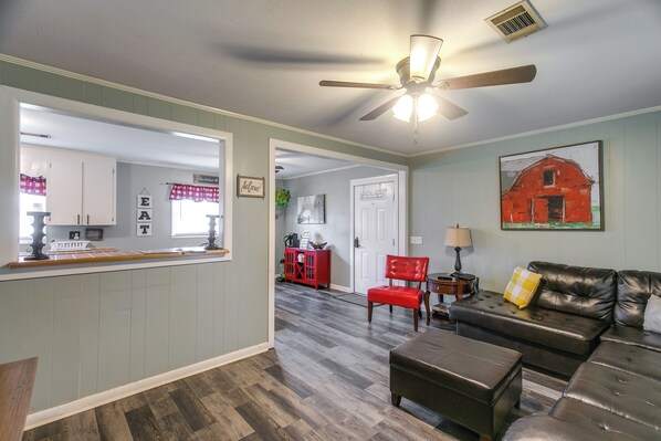 Una sala de estar con un sofá, un reposapiés y una silla roja. Hay una zona de cocina con armarios y un refrigerador.