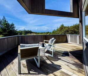 Een houten terras met een bank en stoelen, een bijzettafeltje en uitzicht op bomen en lucht.