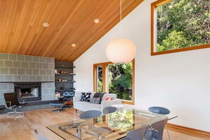 A modern living room with a fireplace, a glass dining table, and a large window with a view of greenery. - Mistral TOT # 4838: 3  BR, 2.5  BA House in The Sea Ranch, Sleeps 6 (The Sea Ranch)