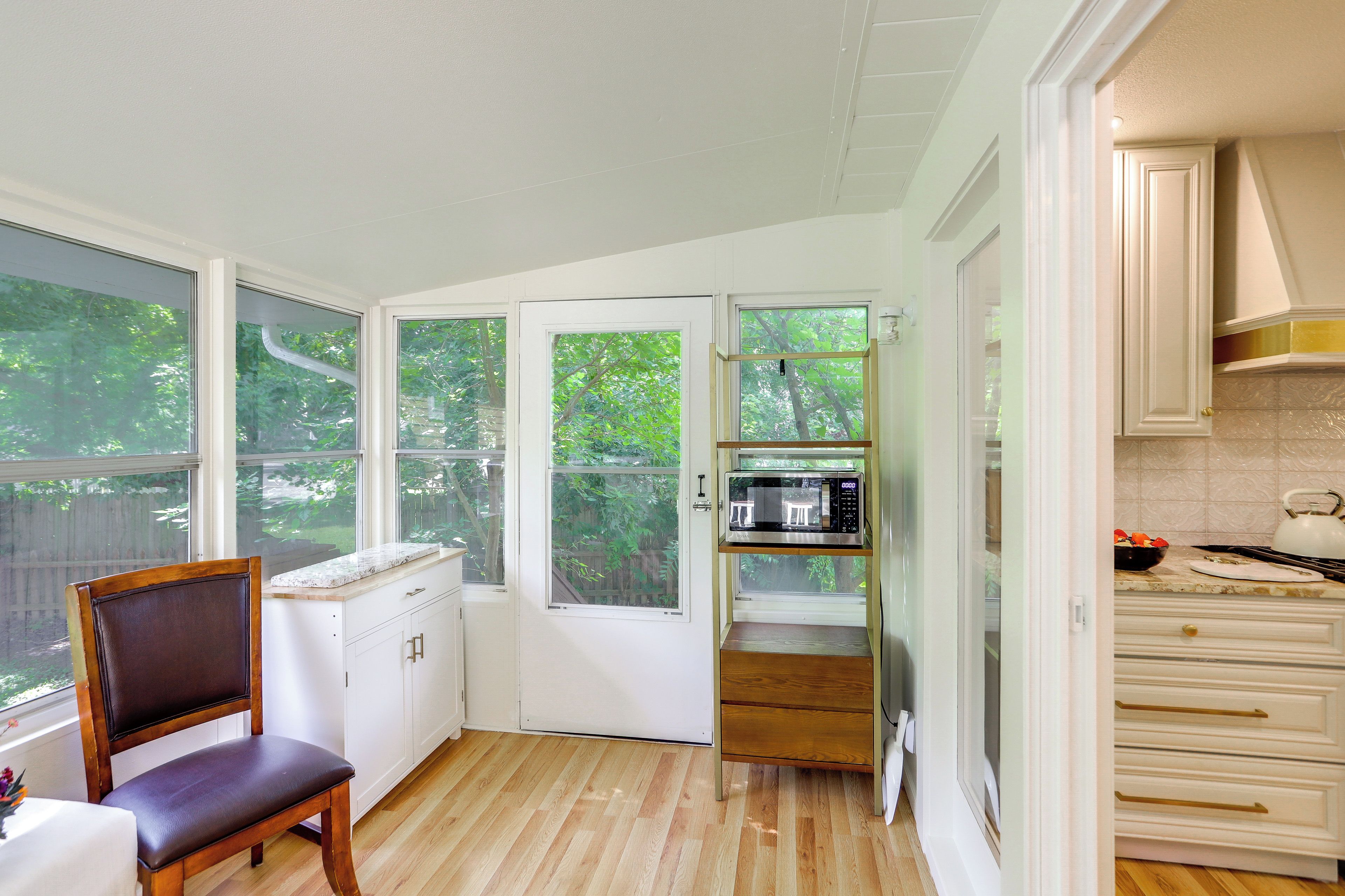 A kitchen with white cabinets, a wooden chair, a microwave, and a view of greenery outside.
