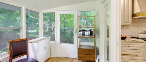 A kitchen with white cabinets, a wooden chair, a microwave, and a view of greenery outside.