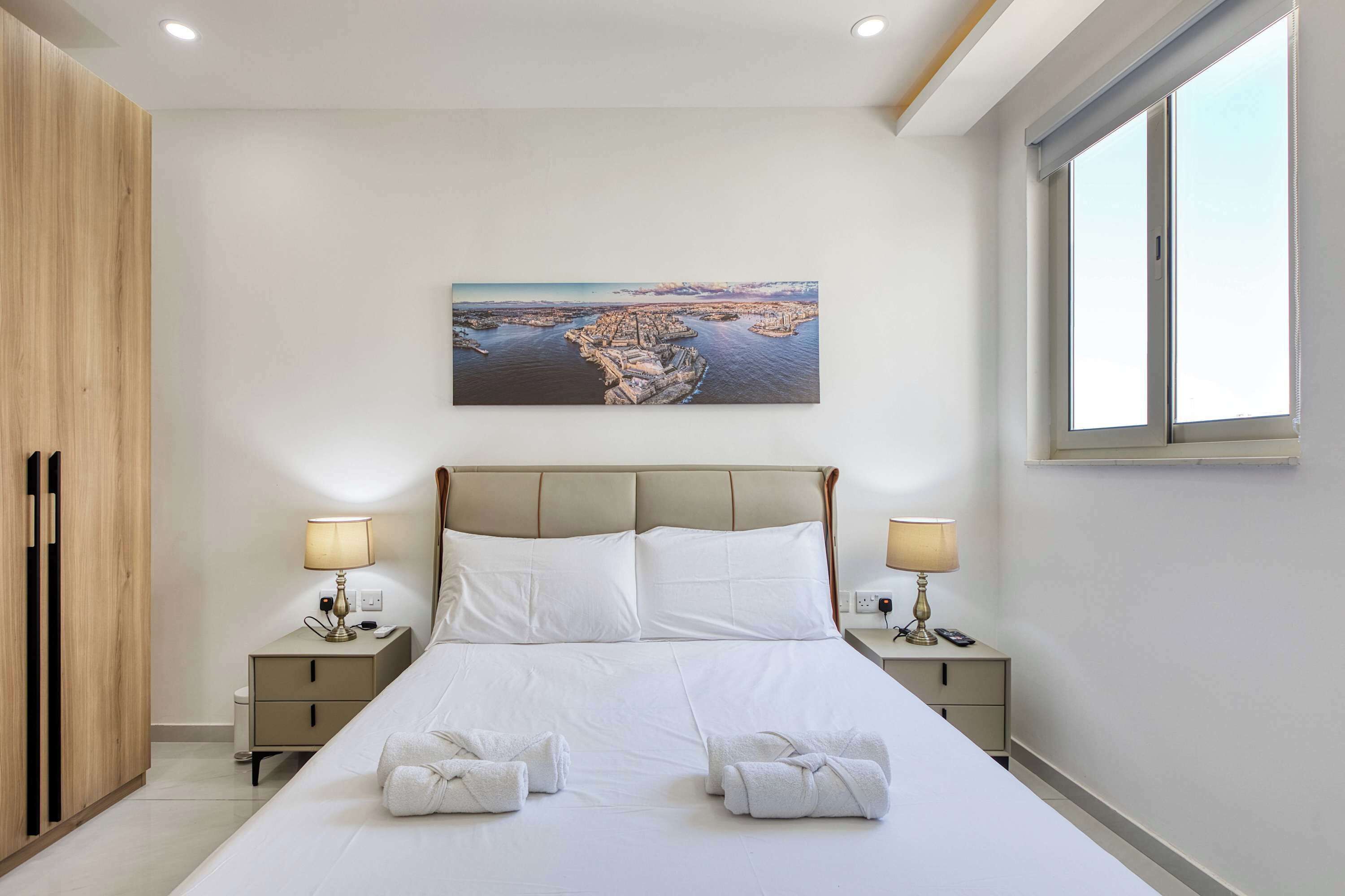 A hotel room with a bed, two bedside tables with lamps, a painting on the wall, and a window.