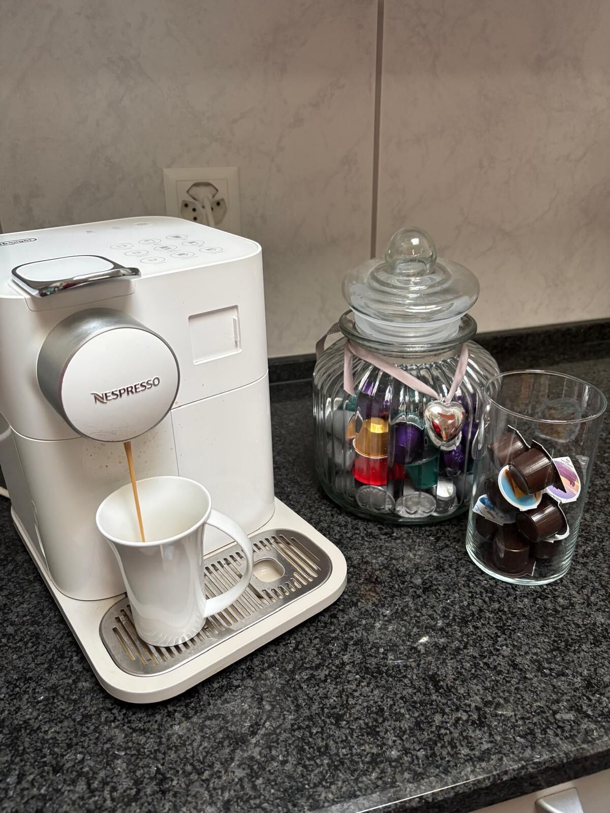 Een witte Nespresso koffiemachine schenkt koffie in een kopje, een glazen pot met kleurrijke objecten, en twee glazen met chocoladebekers op een granieten aanrecht.