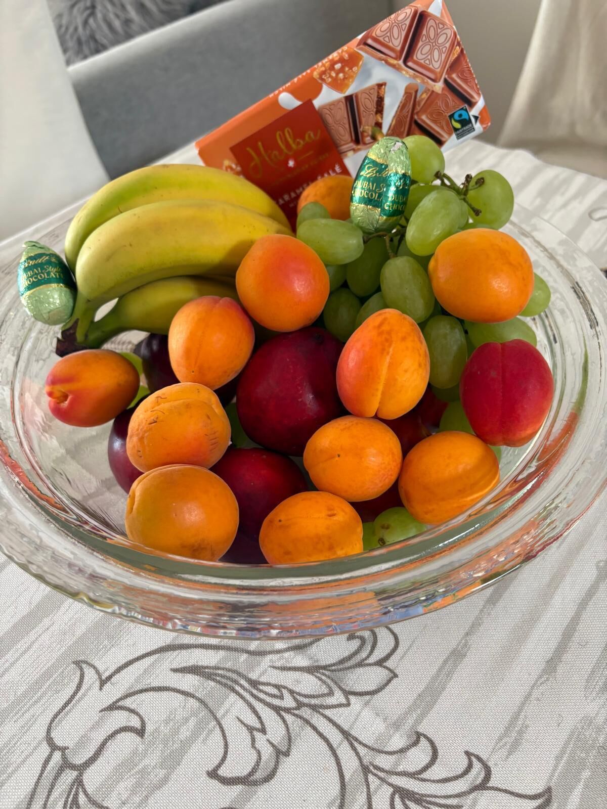 En glasskål fylld med olika sorters frukt och en chokladkaka på ett mönstrat bordsduksunderlägg.