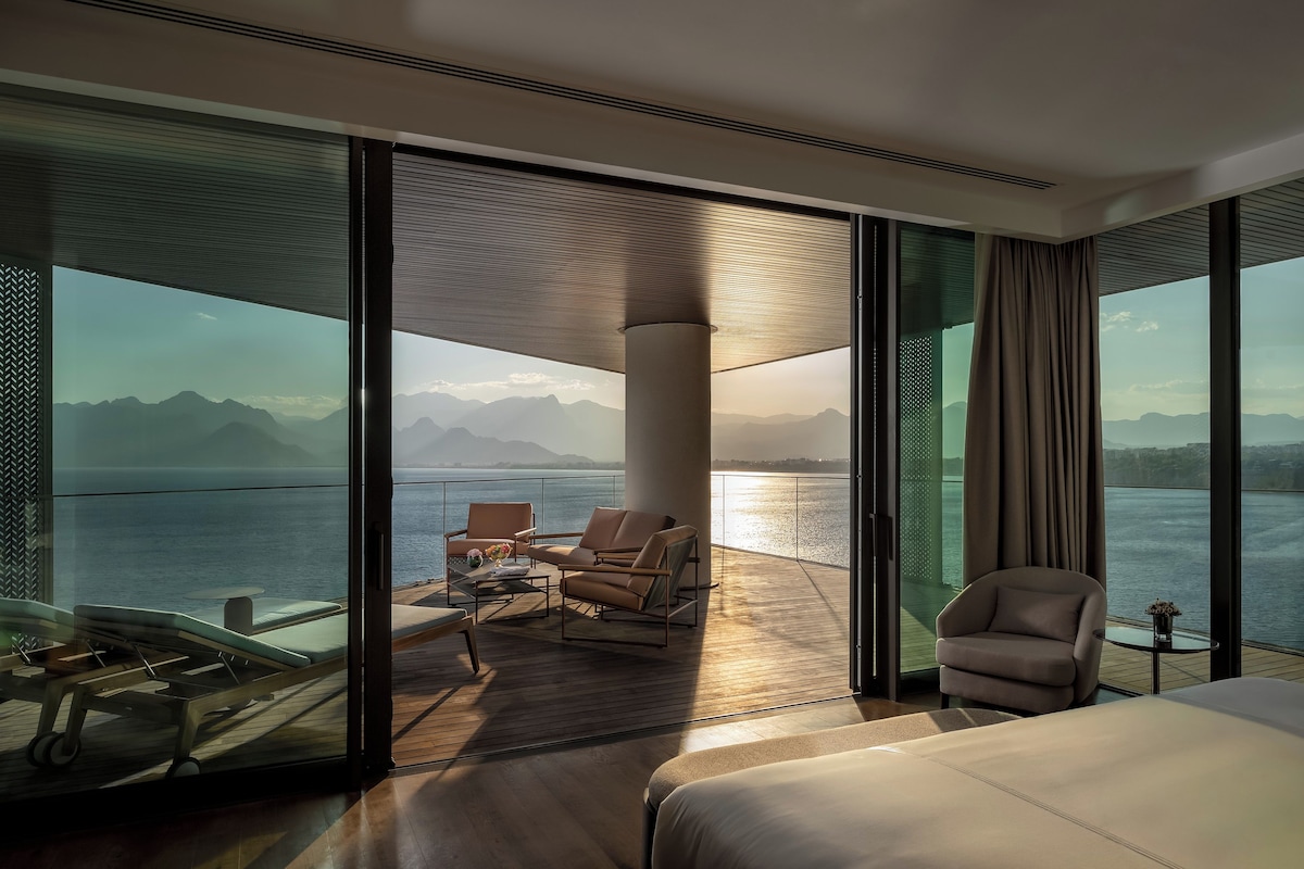 A modern hotel room with a balcony, a bed, and seating area with a view of the sea and mountains.