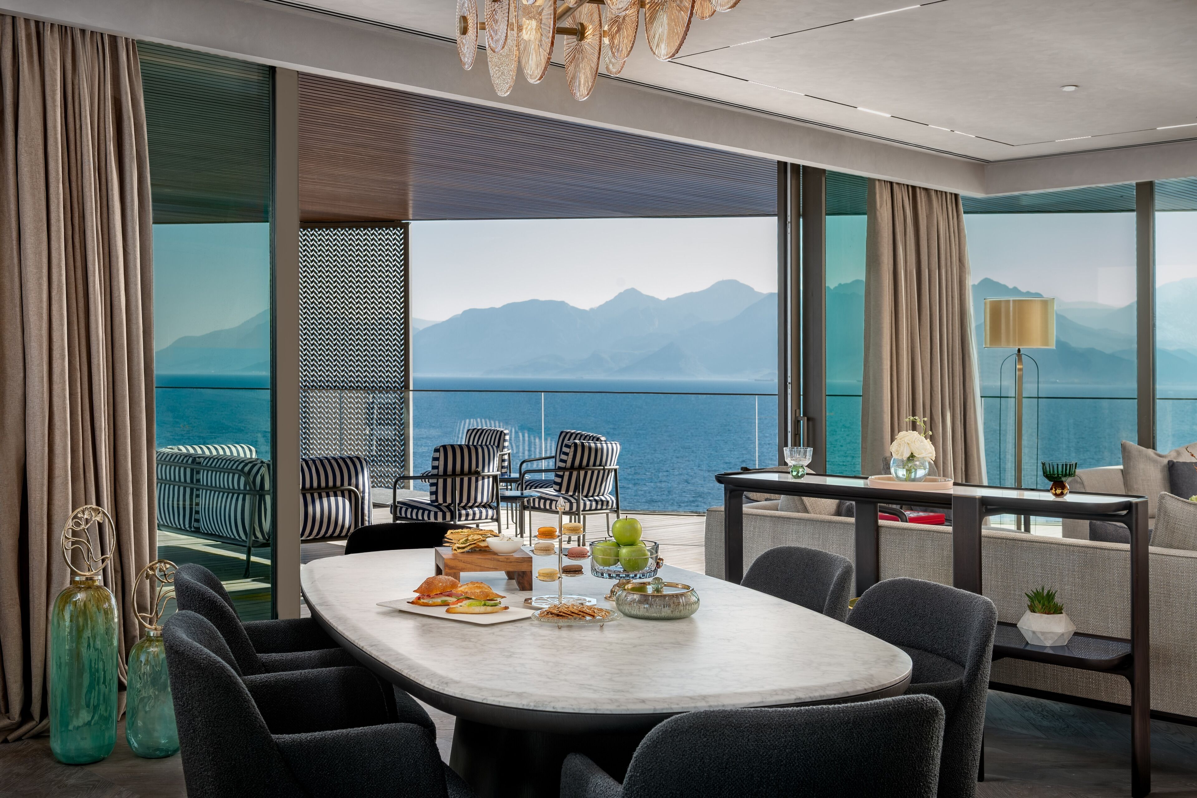 Un espace repas moderne offrant une vue sur la mer et les montagnes, avec une table ronde, des chaises à rayures et une variété de plats et de boissons.