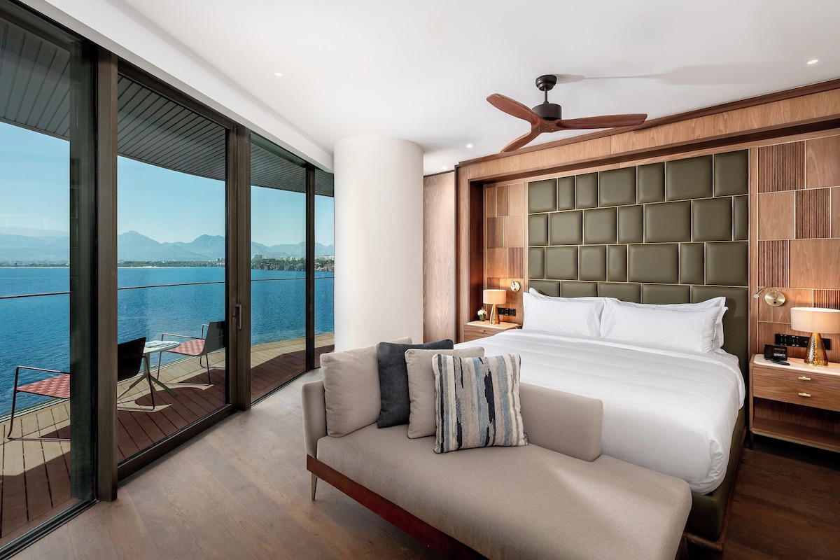 A hotel room with a large bed, a balcony with a view of the sea, and a ceiling fan.
