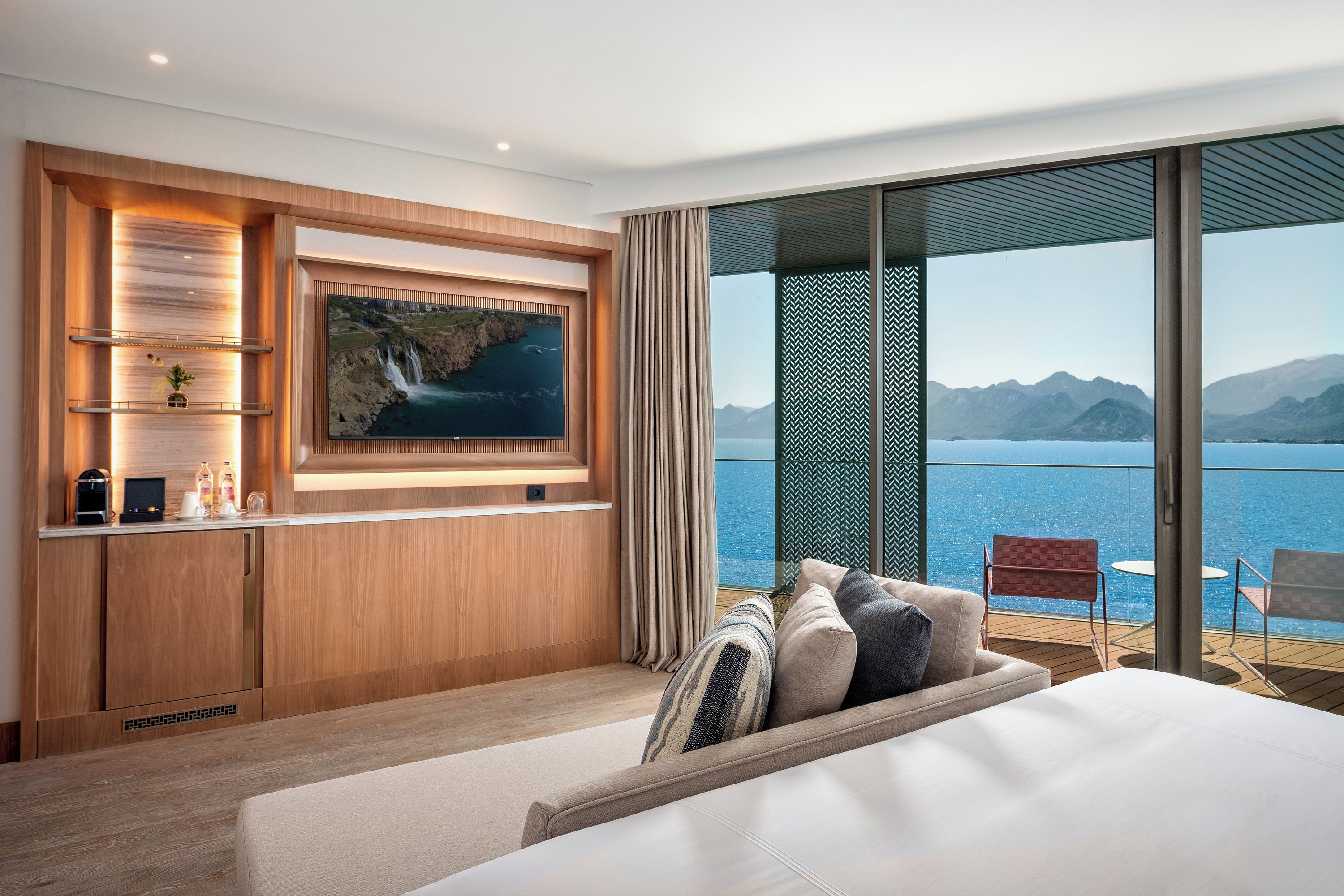 Ett hotellrum med en stor säng, en tv som är monterad på en träpanel, en minibar och en balkong med utsikt över havet och bergen.