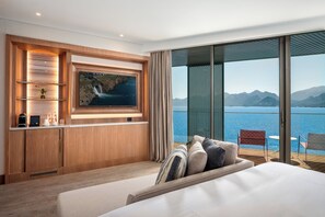 Et hotelværelse med en stor seng, et tv monteret på en træpanel, minibar samt en balkon med udsigt over havet og bjergene.