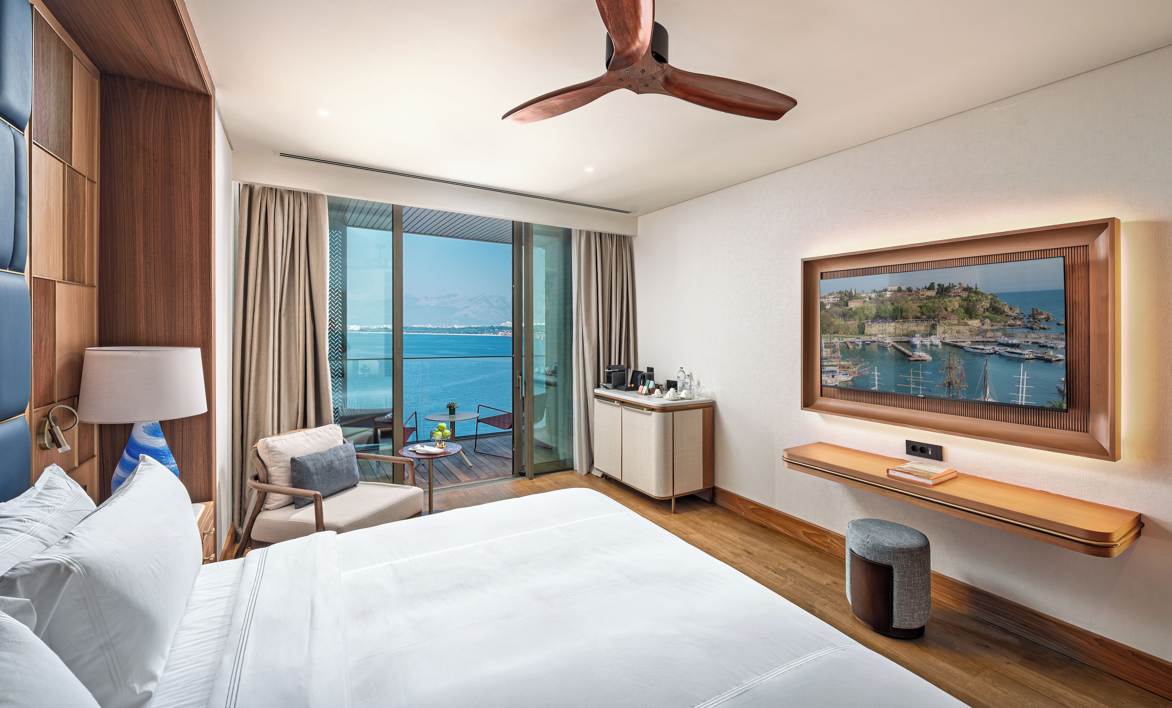 A hotel room with a large bed, a desk with a computer, a chair, a view of the sea, and a ceiling fan.