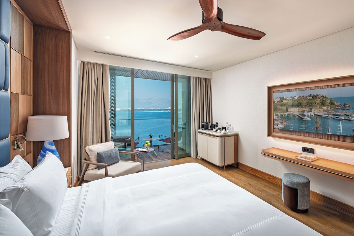 A hotel room with a large bed, a desk with a computer, a chair, a view of the sea, and a ceiling fan.