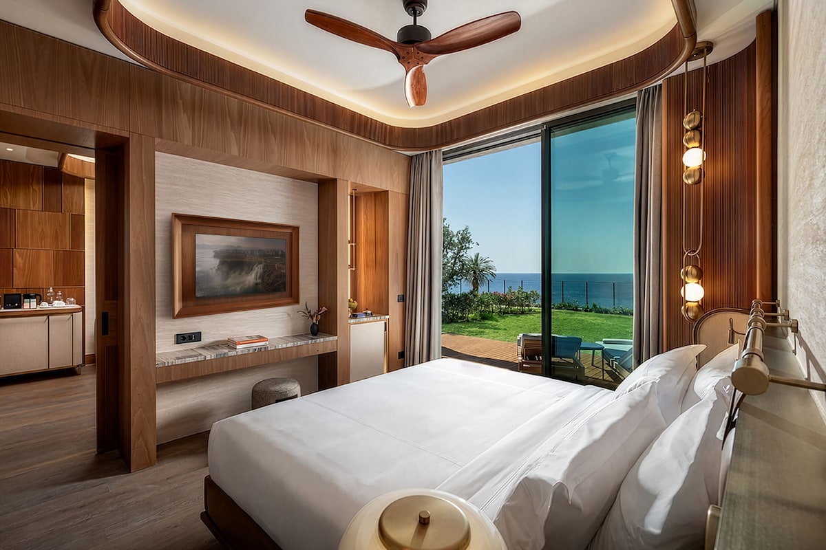 A hotel room with a bed, a desk, a television, and a view of the sea.