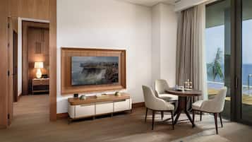 Habitación de hotel con piso de madera, televisor montado en la pared, una mesa de comedor con sillas y vista al océano a través de amplias ventanas.
