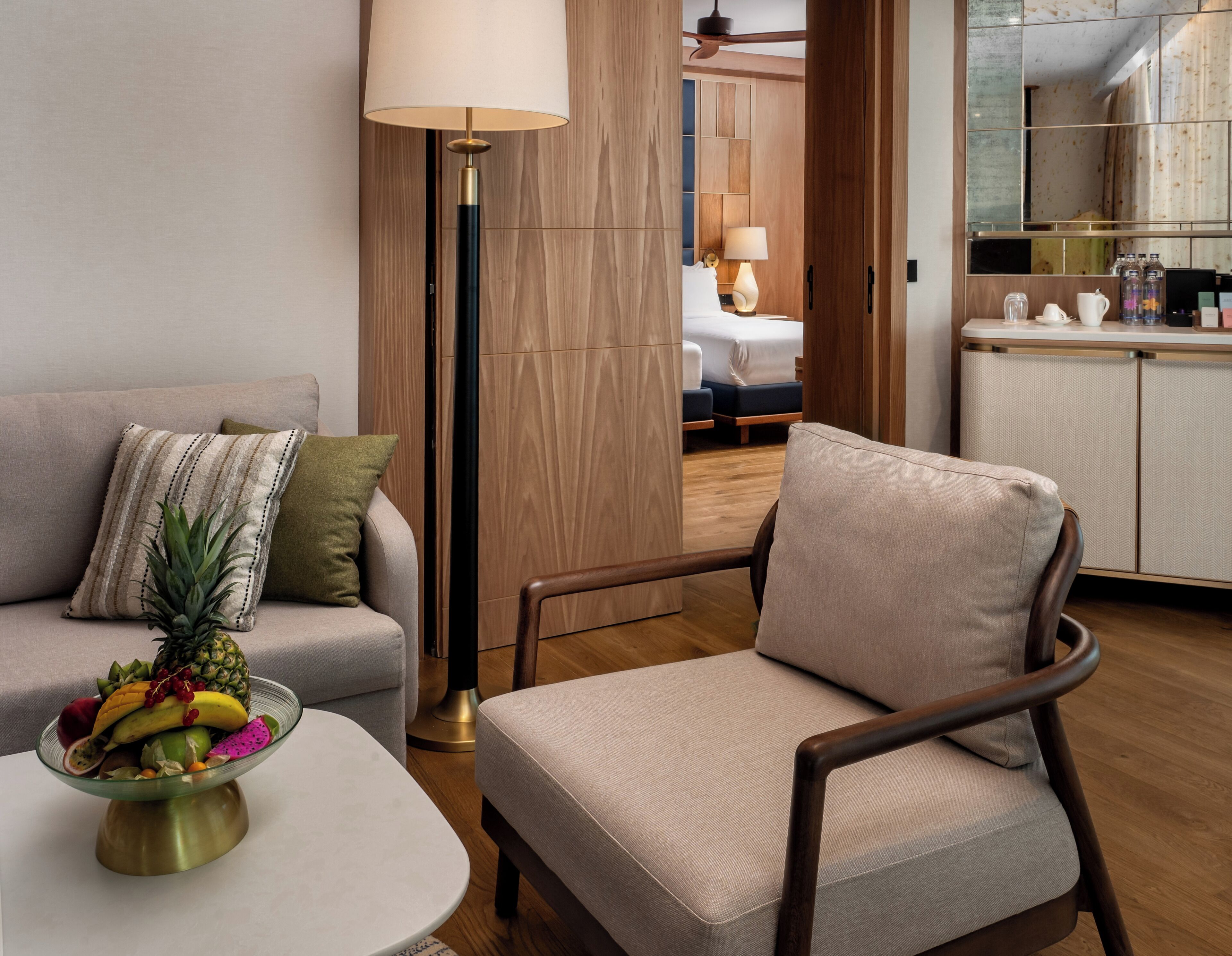 A modern hotel room with a sofa, armchair, and a dining table with fruit.