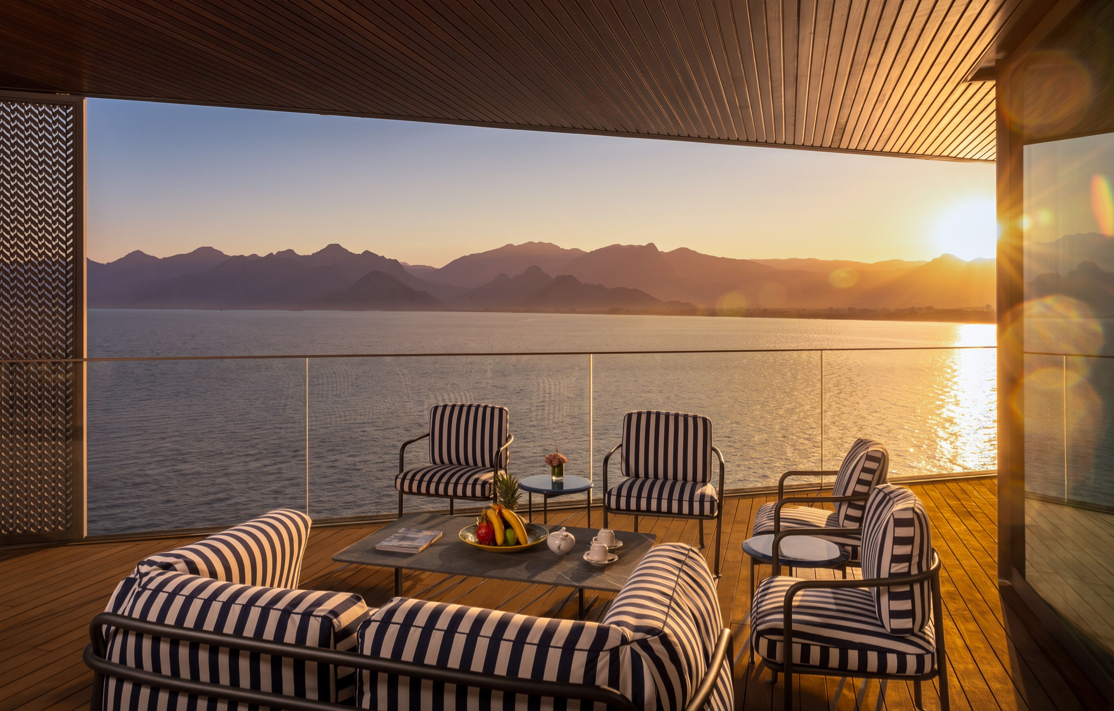 A deck with striped sofas and chairs, a table with a fruit bowl and teapot, and a view of mountains and water at sunset.