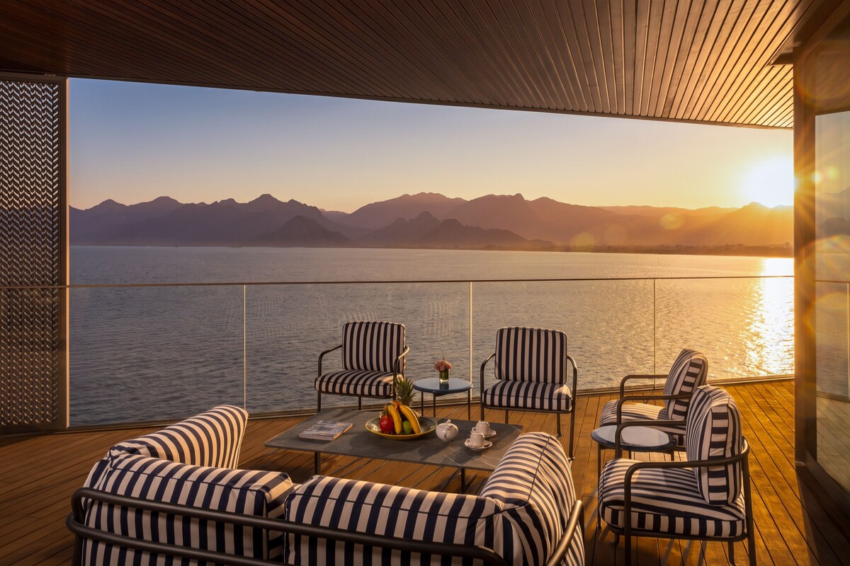 A deck with striped sofas and chairs, a table with a fruit bowl and teapot, and a view of mountains and water at sunset.