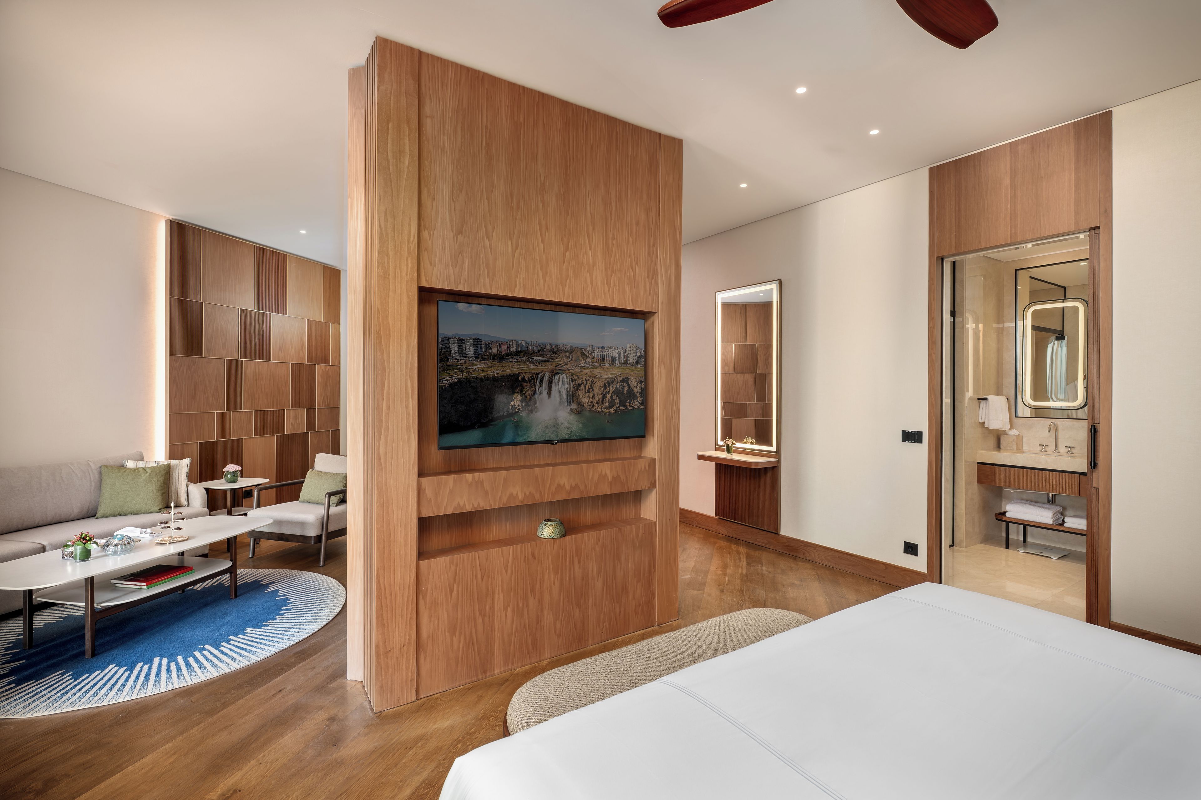 A modern hotel room with a large bed, a TV mounted on a wooden panel, a sofa, a coffee table, and a bathroom with a mirror and sink.