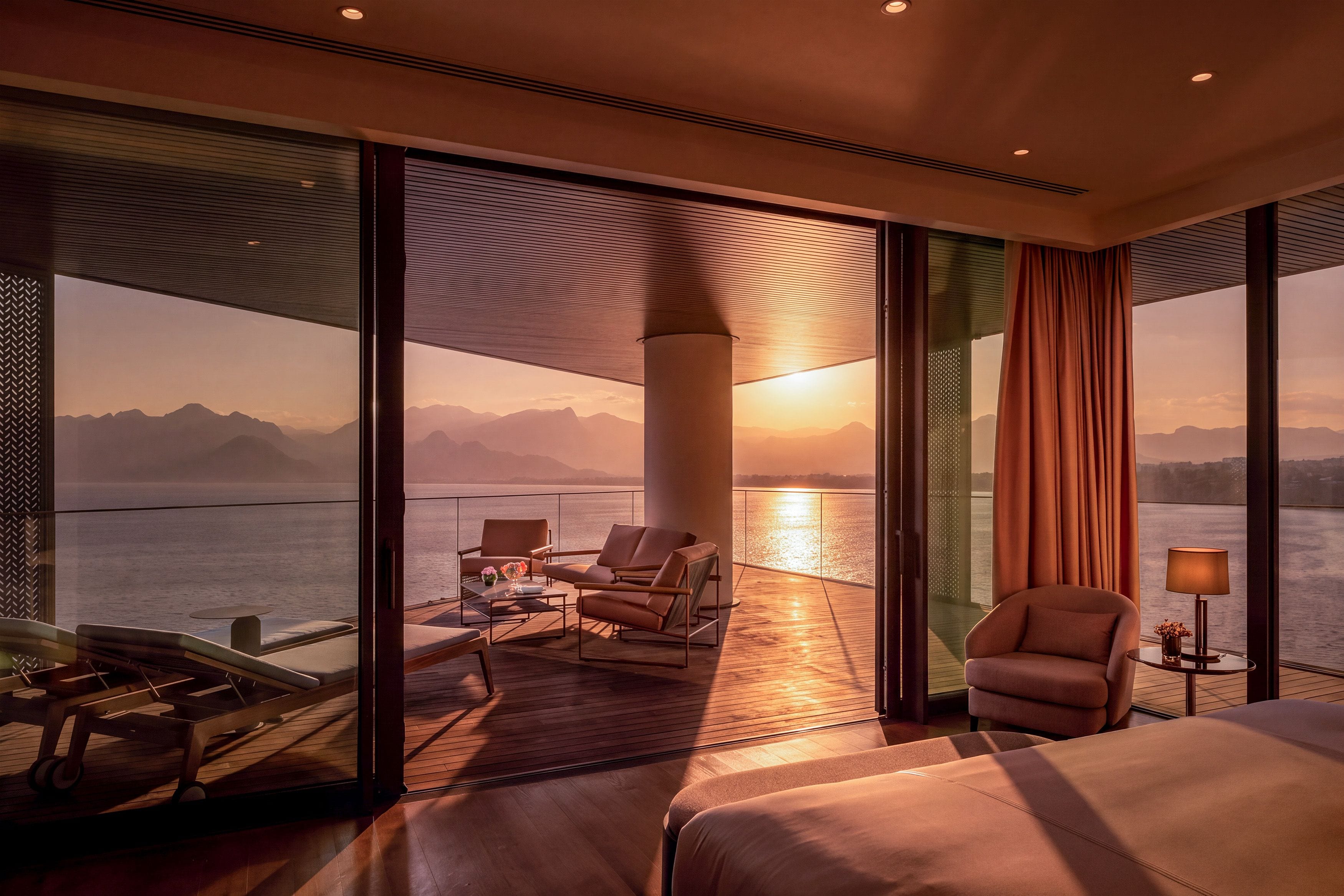 A modern hotel room with a large bed, a seating area with a table, and a view of the sea and mountains.