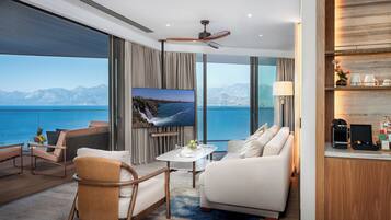 A modern hotel room with a large window offering a view of the sea and mountains, a flat-screen TV, a ceiling fan, and a cozy seating area with a coffee machine.