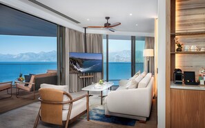 A modern hotel room with a large window offering a view of the sea and mountains, a flat-screen TV, a ceiling fan, and a cozy seating area with a coffee machine. - Divan Talya (Antalya)