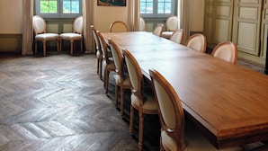 Une salle de conférence avec une grande table en bois, de nombreuses chaises, un tableau accroché au mur et de grandes fenêtres dotées de rideaux.