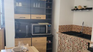 A compact kitchen with a microwave, a dining table set with plates and glasses, a wooden cabinet, and a patterned curtain. - Studio in the Drôme Provençale. Suze's Cocoon. (Suze-la-Rousse)