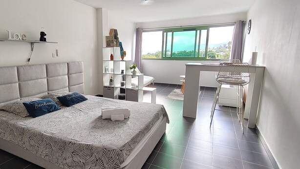 Un dormitorio con cama, escritorio y silla, ventana con vistas, y estantes con objetos decorativos.