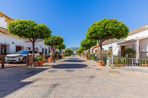 A paved street lined with trees and white buildings, a parked car, and a clear blue sky. - Coral8 - Top modern townhouse in Estepona old town (Estepona)