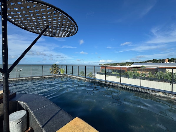 An infinity pool with a view of the ocean, a metal railing, and a lounge area with a table and chairs. - Setai Edition Suites (João Pessoa)