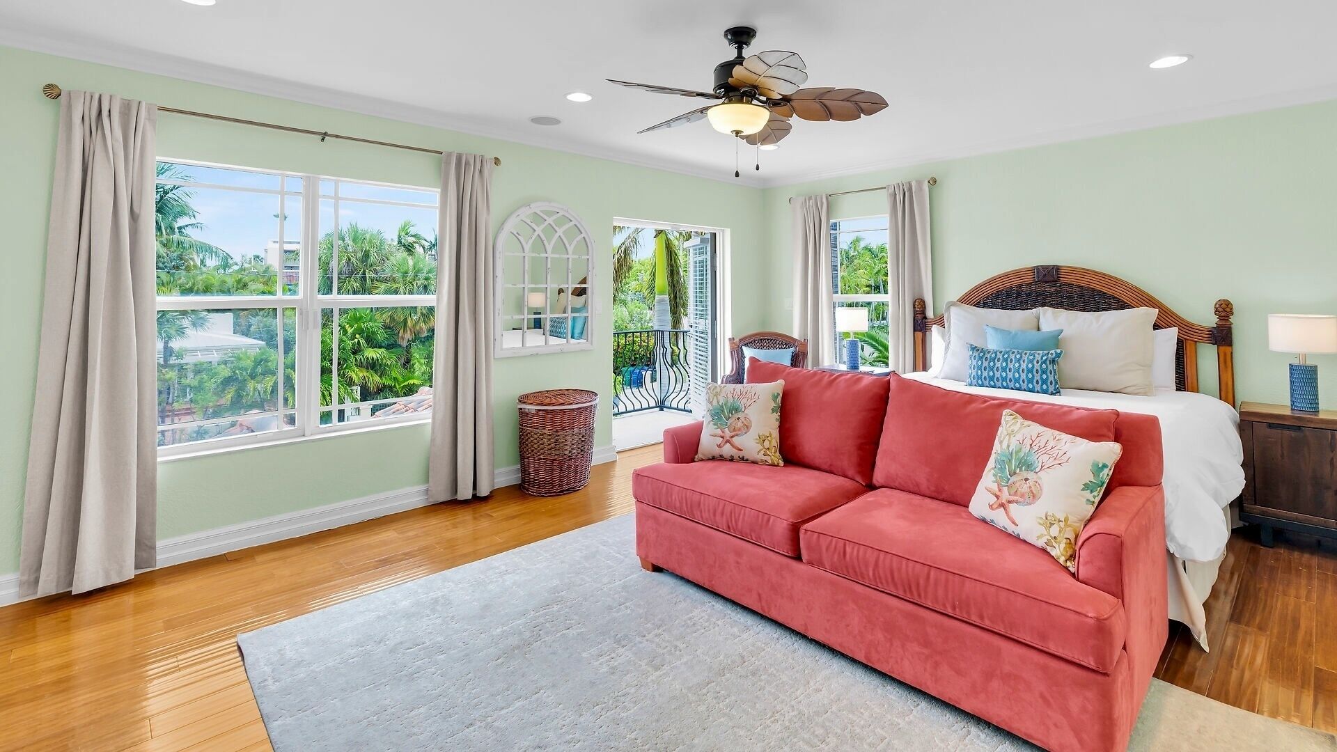 A room with a bed, a red sofa, a ceiling fan, and large windows.