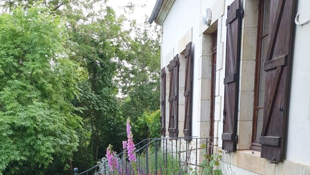 A stone pathway leading to a white building with dark shutters, surrounded by lush greenery and flowering plants. - CHAMBERAT COUNTRY HOUSE (Chambérat)