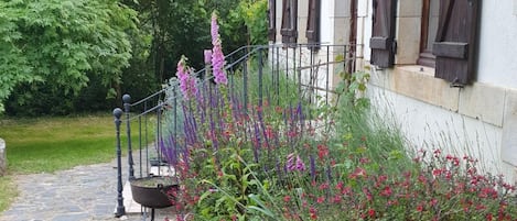 Ein gepflasterter Weg führt zu einem weißen Gebäude mit dunklen Fensterläden, umgeben von üppiger Vegetation und blühenden Pflanzen.