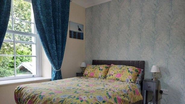 Ein Schlafzimmer mit einer blumigen Tagesdecke, einem Fenster mit Vorhängen, einem Nachttisch mit Lampe und einer Wand mit floralem Wandpapier.