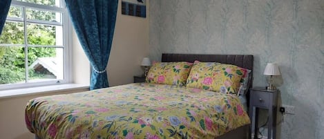 A bedroom with a floral bedspread, a window with curtains, a bedside table with a lamp, and a wall with a floral wallpaper pattern.