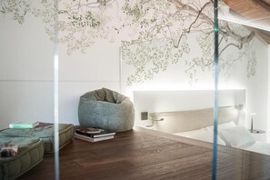A modern bedroom with a wooden floor, a bean bag chair, a bedside table with books, a wall-mounted lamp, and a large window.