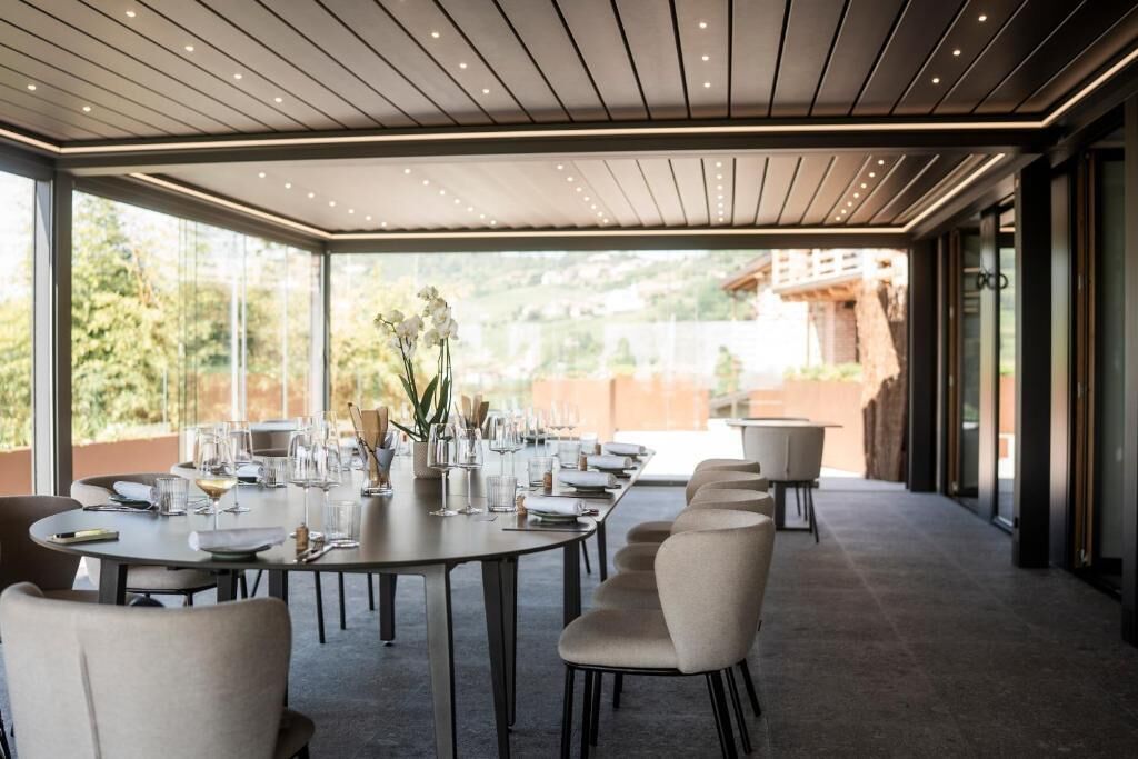 A modern dining area with a long table set for a meal, surrounded by chairs, and large windows offering a view of greenery and a building.