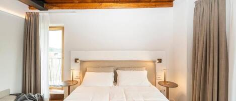 A bedroom with a wooden ceiling, a large bed with white bedding, two bedside tables, and a view of the outdoors through a window.