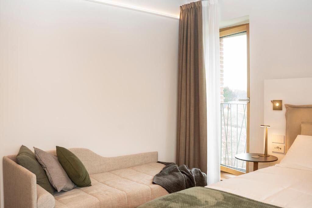 Hotellihuone, jossa on sohva, sänky, pieni pöytä lampun kanssa ja ikkuna, jossa on verhot.