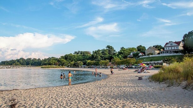 A beach with people swimming and sunbathing, a sandy shore, and buildings in the background.