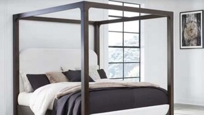 A four-poster bed with a dark frame and white bedding in a room with a large window and a framed picture on the wall.