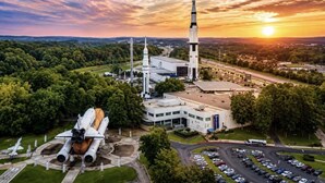 A space shuttle on display, a tall rocket structure, and a building with a parking lot.
